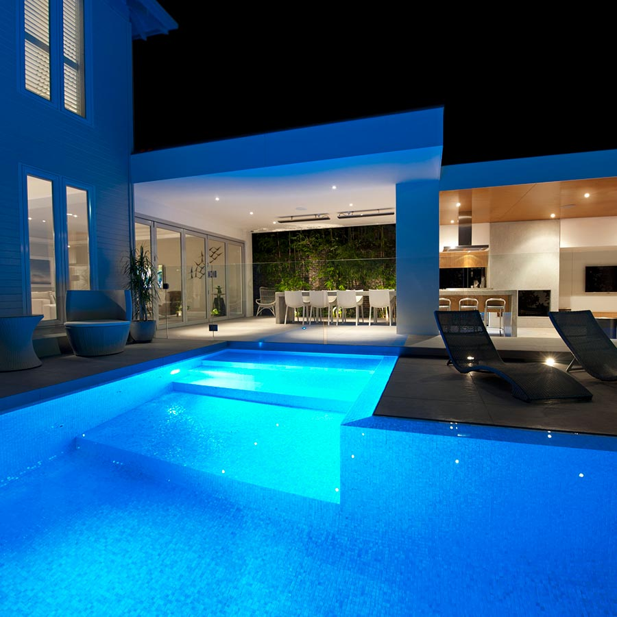 Modern backyard pool lit bright blue at night beside a patio and open living area